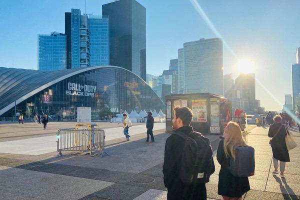 A sunny, wide shot of a modern city square with people walking. A curved building with a "Call of Duty Black Ops" banner is prominent in the center.